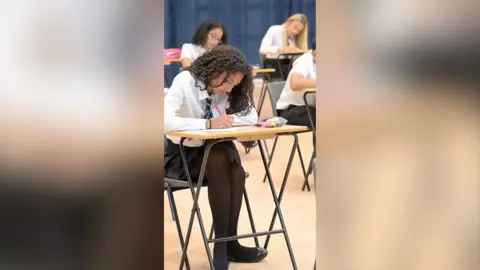 Teenagers sit at desks writing in an exam hall.