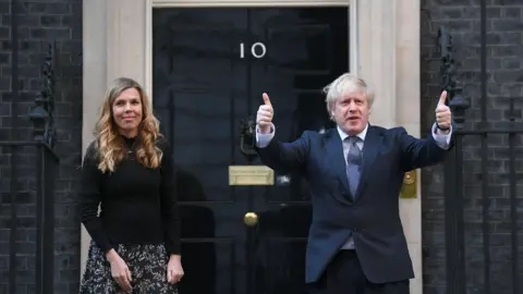 Getty Images Carrie Johnson stands outside 10 Downing St with her husband, former prime minister Boris Johnson, who is doing a thumbs-up gesture.