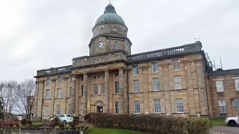 An exterior shot of Dr Gray's on a grey cloudy day, the sandstone building with four columns rises up to a copper dome above an old clock face.