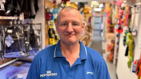 A man wearing glasses and a blue polo shirt, with a white logo on the right side, and 'The Pet Pantry' written in white letters on the left side, stand in a pet shop. Pet toys and other items can be seen in the background. 