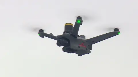 An airborne drone with green lights on all three visible propellor arms. 