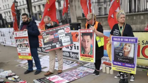 Inquest charity Families in London protesting at the number of mental health deaths in Essex. There are two men and two women holding placards with pictures of their loved ones, alongside words including 