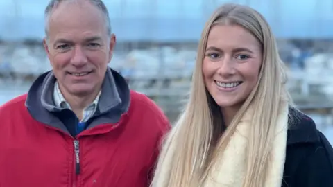 Ella Dawson Foundation Adam Waugh and Natasha Dawson. They are posing together and smiling at the camera. Mr Waugh has grey short hair and is wearing a red jacket. Natasha Dawson has long blond hair and is wearing a white scarf and a black coat.