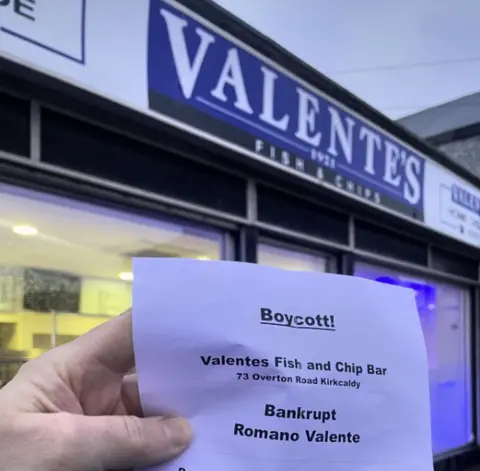 Valentes Close up of a hand holding a white paper flyer calling for a boycott of Valentes Fish and Chip Bar. 