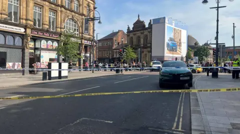 A road with a yellow police cordon around it. There is a car in the middle of the road with police cars and officers in the background. There are shops surrounding the road and crowds of people on the roadside.