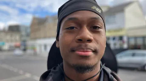 Kaleb, 19, from Tottenham is wearing a black hat which covers the back of his head. He is in a street which has been blurred to focus on his face.