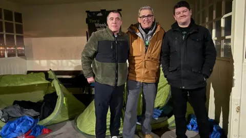 Sanctuary Trust Tim Ringsdore is smiling at the camera and has his arms around Deputy Sam Mezec and support worker Sean Culkin. It is night-time and they are standing in a building that looks like a bus shelter that has windows. There are tents pitched on the concrete floor which have sleeping bags in them.