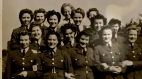 Cynthia Evans Cynthia Evans is in a black and white photograph with 14 of her female RAF colleagues. They are all wearing their RAF uniforms and are smiling at the camera.