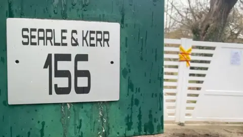 There is a tear color box that says in the foreground "Searle & Kerr 156". The bright yellow and bright red police tape covers a white door in the background.