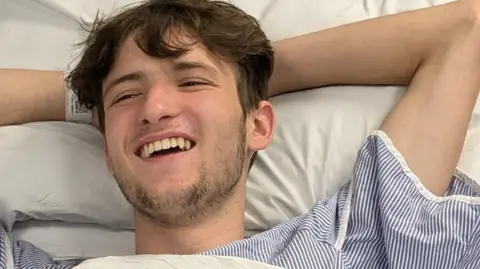 Family Photo Arlo is lying in a hospital bed, with his hands behind his head in a relaxed position and he smiles looking off camera. He wears a blue and whit epinstripe hospital gown and the bedding he is in is white. He has shorter brown hair and some stubble lining his jaw and chin.