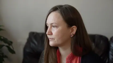 Martyna stares out of the window sadly. She has long dark hair and wears an orange top and black cardigan. 