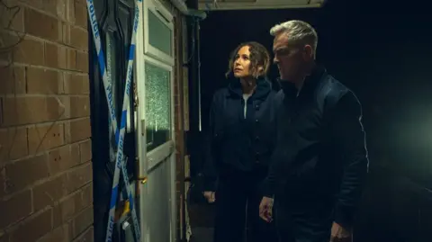 Run Away Simon Greene, played by James Nesbitt, is standing in front of a door which has police tape across it. His wife Ingrid Greene, played by Minnie Driver, is standing next to him. They both have concerned looks on their faces. 