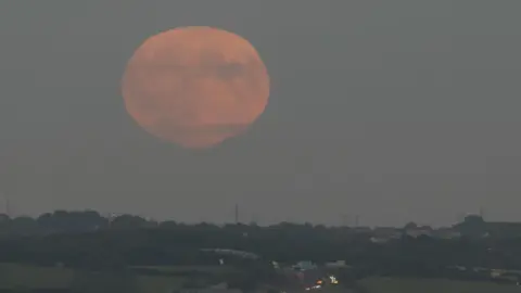 BBC Weather Watchers/Andy Denison A coppery supermoon above Armley in Leeds. The city's skyline can be seen in the distance behind green hills and trees.