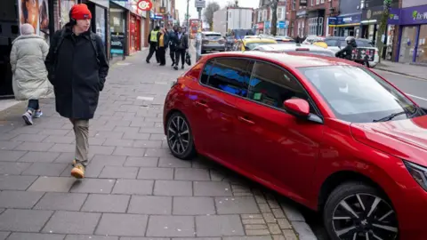 A red car parked blocking the pavement with people looking at it, walking by.