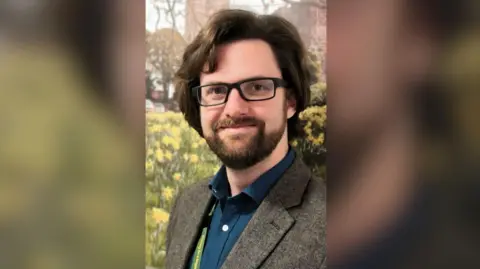 A bearded man with dark, long brown hair and black-framed glasses He is wearing a grey suit and a teal shirt and standing in front of a church tower and a patch of daffodils. 