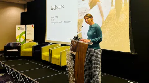 Jersey Community Foundation A woman standing on stage behind the podium speaks into a microphone behind which is a power supply showing a welcome screen. She is wearing a blue long-sleeve top and black and white checkered wide leg pants.