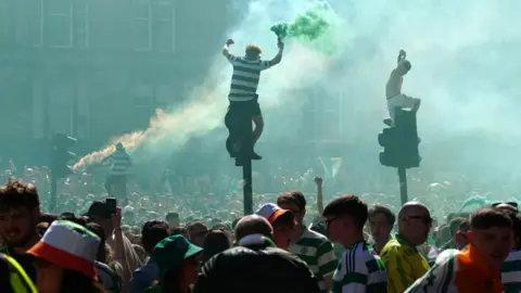 Celtic fans sitting top of traffic lights with hundreds of other fans on the street below them. One fan on a traffic light is holding a smoke bomb and there is more smoke at the back of the photo  