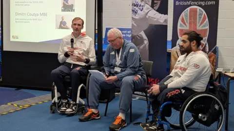 Two wheelchair fencers wearing their Team GB training kit are sat either side of a man holding a microphone. One of them, Paralympic champion Piers Gilliver, is holding paper and also holding a microphone.