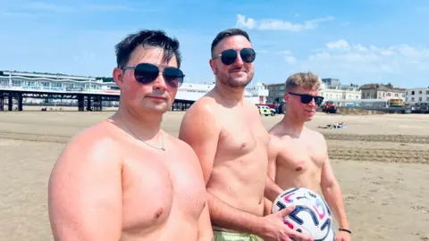 Three topless men on Weston beach. One of the men is holding a white, blue and red football. They are all wearing sunglasses.