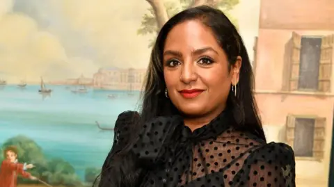 Anita Bhagwandas Anita smiling whist looking at camera. She wears a black mesh dress with a bow on the left corner. Anita has dark brown hair. 