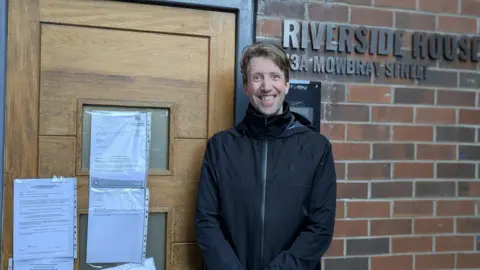 Peter Jones A man with light brown hair is wearing a black zipped up jacket and smiling. He is standing by a brick wall and a wooden door with several pieces of paper pinned to it. There are silver letters on the wall which say Riverside House