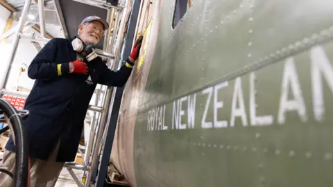 Aerospace Bristol A volunteer standing beside the aircraft, resting one hand on its side. The older man is wearing a long navy work coat, red gloves and a breathing ventilator. On the side of the plane it reads 'Royal New Zealand' before the words are cropped out.