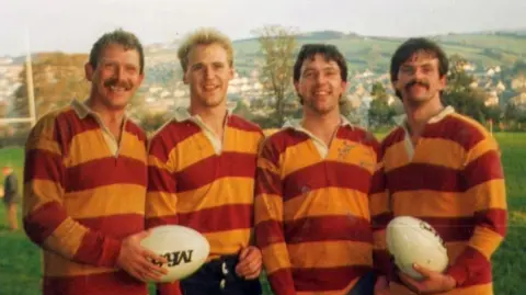 Rob Ewen Rob Ewen stood on the left with his three brothers. They are wearing red and orange striped rugby tops and two of them are holding rugby balls. 