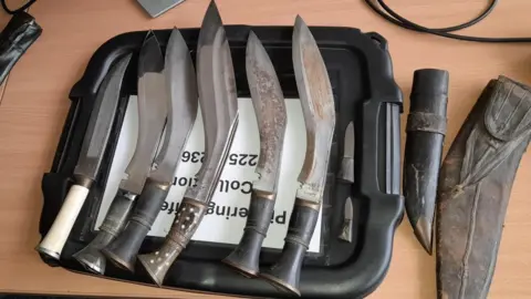 Five large knives and two smaller blades on top of the lid of a plastic box, on a desk in an office.