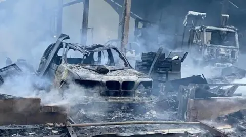 A burnt-out vehicles, with smoke rising. The majority of the image shows a burnt out car, its frame buckled, behind it can be seen another vehicle. On the far right is a burnt out lorry. There is lots of other debris. 