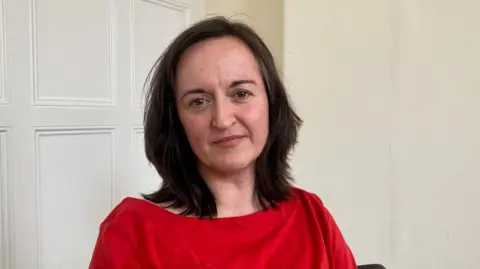 A woman with dark hair and wearing a red top sitting down in front of a white wooden panelled wall