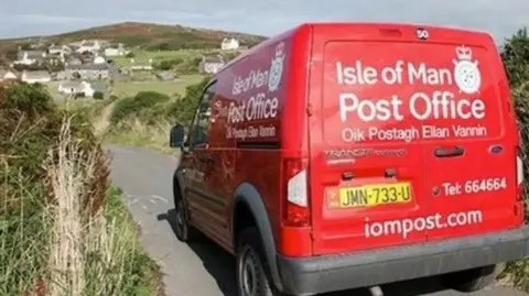 IOMPO A red postal van with Isle of Man Post Office in white on the back parked on a country lane. A group of houses can be seen in the distance.