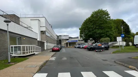 A general view of a hospital entrance. Cars are parked outside of the hospital. There are trees located on land in front of the hospital building.