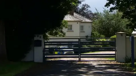 The outside of a gated house. A police car can be seen on the driveway