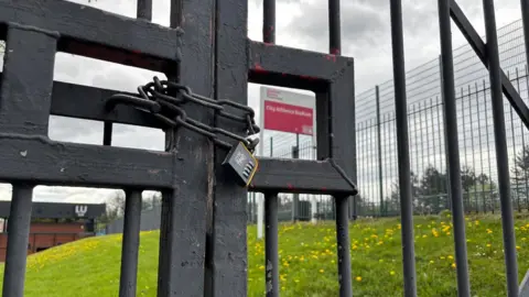 Some large black metal gates locked by a huge padlock. On the other side of the gates are a grassy bank and a pink sign