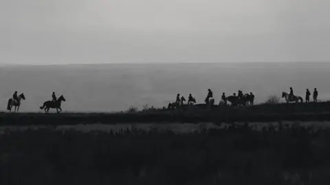 A achromatic  and achromatic  photograph  of a hunt, silhouetted against the sky