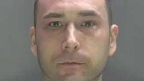 A mugshot of Jason Higgins who has very short brown hair and is wearing a grey top. He has a blank expression on his face. The wall behind him is also grey.