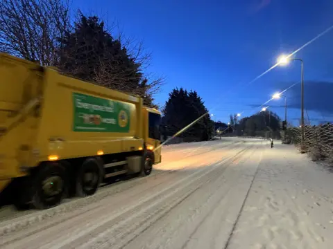 A bin lorry travelling along a snow-covered road in Inverness in the early hours - the sky is still dark though the sun is rising