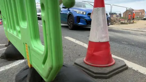 A blue car passes a construction site with two workers in orange jackets. In the foreground are a traffic cone and a green plastic barrier.
