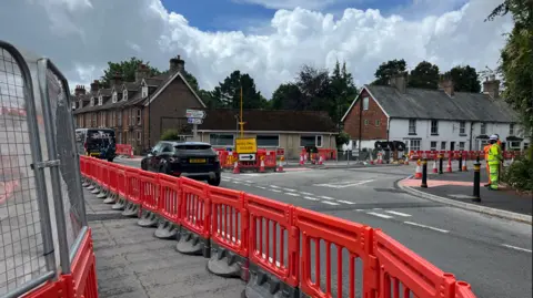 George Carden/BBC Orange plastic fencing and cones placed next to the road with cars driving past, there is also a new pavement where two workers stand
