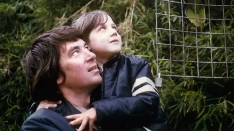 Supplied An image of a primary-school age Helen MacDonald being held by her dad. They are standing in front of trees and looking up at the sky. 