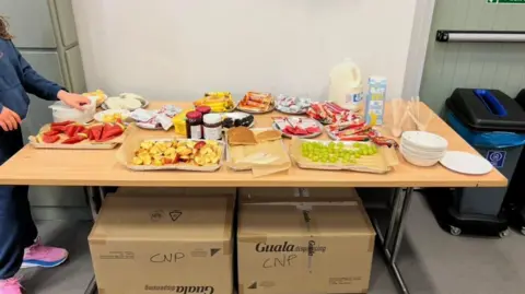 A table with different types of food. Food on the table includes water melon, peaches, grapes, milk, pancakes and different spreads like jam and nutella. 