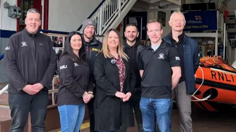 Seven people, five men and one woman stand in a lifeboat shed looking at the camera. A small lifeboat is in the background