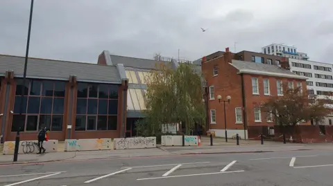 A picture of Eaton Court, a modern three-storey building on the side of a main road, with large concrete blocks placed outside it, with bollards standing in front of another part of it. The Royal Metrological Society, a three-storey Georgian townhouse building, is to the right of the picture. 