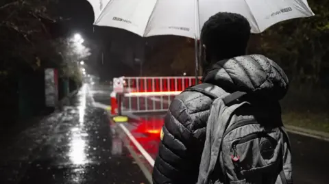 Mr Morakinyo looks over at the gates of Campsfield on a dark, rainy evening.