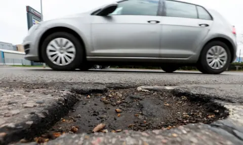 Reuters A pothole is in close-up at the bottom of the frame, as a silver four-door car with silver alloy wheels, passes it on a road.