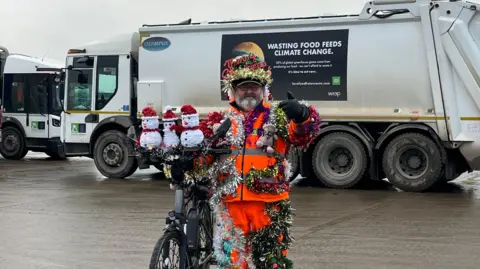 Tom Jackson/BBC Lenny Edward stood in front of a bin lorry with a bike. He has decorated his bike with three white snowmen in red Santa hats. They are sat on top of his handlebars. 