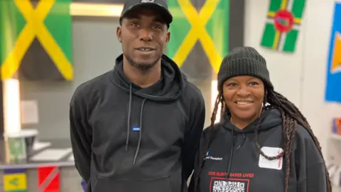 A man and woman stand next to each other with Caribbean flags behind