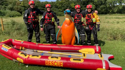 BBC Four firefighters in black and red water rescue gear stand with a tall blue and orange penguin sculpture between them, in front of them is a red inflatable dinghy that has the word rescue written on the side
