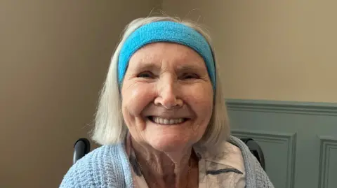 Steve Jones/BBC. Yvonne Noble, una anciana que lleva un cárdigan azul claro y una diadema azul. En la foto aparece sonriendo.