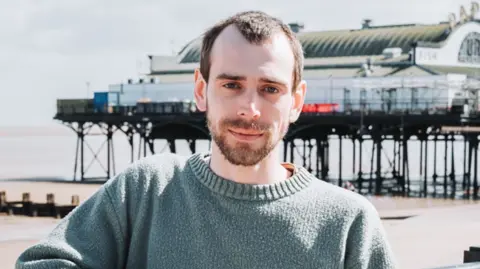 Green Party candidate Leon Brown he has short brown hair and beard and is wearing a green jumper and is standing in front of Cleethorpes Pier